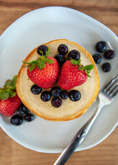 A stack of pancakes on a plate, topped with strawberries and blueberries, with more berries scattered along the plate with a fork.