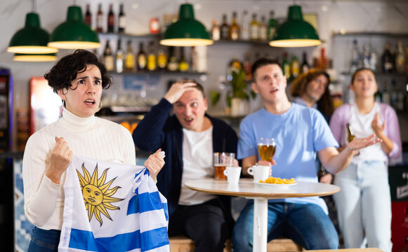 Emotional sad young female fan supporting team and holding state flag of Uruguay in hands during rest in sports bar