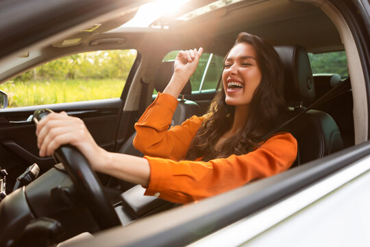 Overjoyed pretty young woman driver listening to music and singing while holding one hand on steering wheel, enjoying auto ride