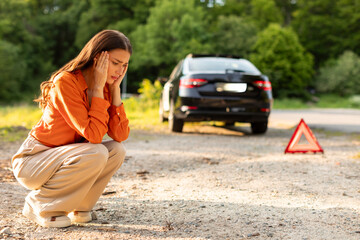 Sad woman driver sitting on roadside with broken car on background and red warning triangle sign, upset lady waiting for help