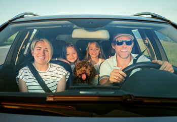 Happy family enjoys road trip together, with fluffy brown Maltipoo dog in front seat and two young...