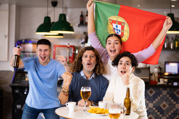 Group of emotional young adult fans supporting Portuguese sports team with state flag while resting...