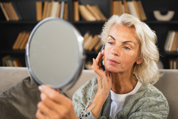Love yourself. Beautiful old mature woman enjoying mirror reflection. Senior adult older lady looking at mirror confident happy on sofa at home. Woman doing daily morning beauty routine. Self love