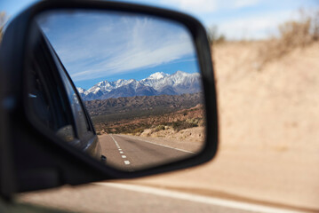 The view of the imposing snowy Andes Mountains in Mendoza, Argentina is reflected in the rear view mirror of a car traveling on the highway.
