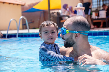 A cheerful father and son are having a great time in the water under the shining sun, smiling as they teach swimming. They look at the camera, capturing a real-life moment that strengthens their bond