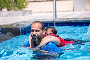 A cheerful father and son are having a great time in the water under the shining sun, smiling as they teach swimming. They look at the camera, capturing a real-life moment that strengthens their bond