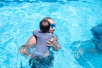 A young father is helping his child begin swimming, teaching him the basics and ensuring he isn't afraid of drowning or getting water in his ears, mouth, and nose