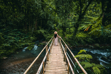 Fototapeta premium The young woman in a dress and hat is walking on the bridge, looking around in a tropical forest. Leke Leke Waterfalll Trail. Travel concept.