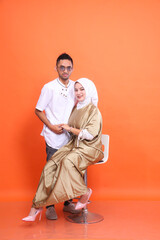 Asian muslim couple (man and woman) smiling holding hands wearing kaftans and muslim clothes in the month of Ramadan isolated orange background. Islamic religious, fashion, commercial concepts