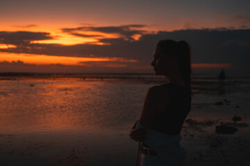 Fototapeta premium Woman contemplating the sunset by the sea, captured in silhouette. The vibrant sky and reflective water create a serene, contemplative scene, emphasizing the beauty and tranquility of the moment.