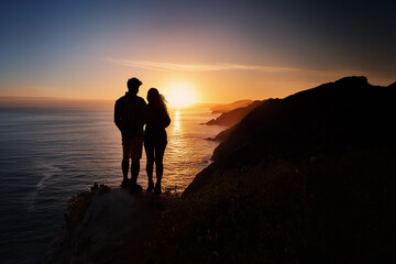 Young romantic couple together at sunset, looking over the ocean as the sun sets below the horizon. Man and woman standing on cliffs in silhouette.