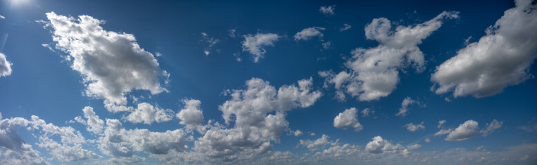 vA panoramic view of only the bright summer blue sky with white clouds. The sun is peaking into the top of the image.
