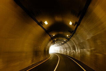 A long tunnel among the mountains. A light in the end of a tunnel. Industrial landscape. Asphalt road in the mountains. Highway. Driving through a tunnel among the mountains on a sunny summer day.