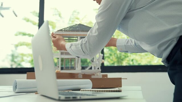 Close up of professional male architect engineer measure house model and using laptop analysis data at meeting table with house model and map . scatter around. Focus on hand. Immaculate.