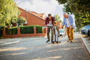 Obraz premium Father and grandfather teaching young boy to ride a scooter on suburban street