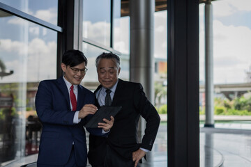 Two men in suits are looking at a tablet. One of them is smiling. Scene is professional and friendly