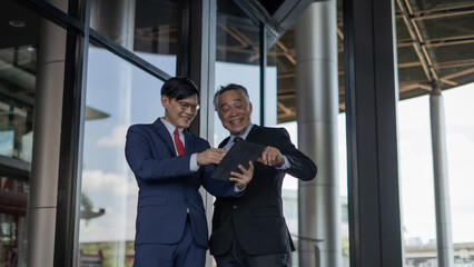 Two men in suits are looking at a tablet. One of them is smiling. Scene is professional and friendly