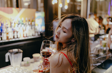 Sexy asian brunette woman tipsy wearing red dress holding a glass of alcoholic drink at the bar. Lifestyle and nightlife concept.