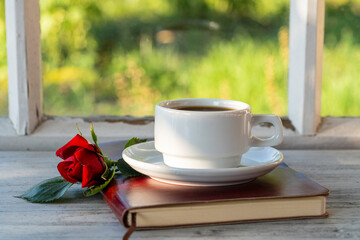 Ceramic white cup of coffee, red rose and notebook on the table in front of the window overlooking the summer garden, closeup