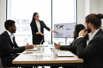 Businesswoman gives report to multiracial colleagues standing near graphs on monitor during office meeting. Diverse team listens to presentation focused on business strategies and data analysis.