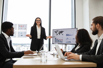 Fototapeta premium Business woman gives report to colleagues standing near graphs on monitor, screen, display, TV. Office setting with city view in background. Professional attire, teamwork, and corporate environment.