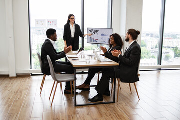 Fototapeta premium Caucasian business woman makes a report to multiracial, multinational, multicultural colleagues standing near monitor with graphs, charts. Diverse business people sit at table and clap their hands.