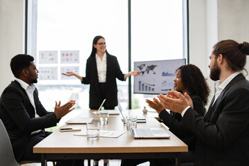 Caucasian business woman makes a report to multiracial, multinational, multicultural colleagues standing near monitor with graphs, charts. Diverse business people sit at table and clap their hands.