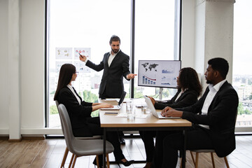 Business man giving presentation to multiracial colleagues standing near graphs and charts on window glass. Team members sitting at table with laptops and participating in discussion.