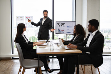 Young businessman providing presentation to multiracial colleagues standing near graphs and charts on window glass. Team sitting at table with laptops engaged in discussion.