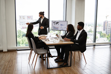 Caucasian business male giving presentation to multiracial colleagues standing near graphs and charts on glass. Office setting with professionally dressed team members paying attention to presenter.