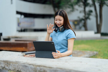 Happy Student Waving at Laptop Screen While Studying Outdoors with Books and Headphones