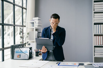 Naklejka premium Businessman in Modern Office Holding Tablet and Looking Stressed with Financial Charts on Desk