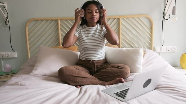 Black woman doing meditation on bed following online wellness program using laptop and headphones. 4k resolution.