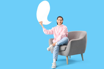 Young woman with blank speech bubble sitting in armchair on blue background