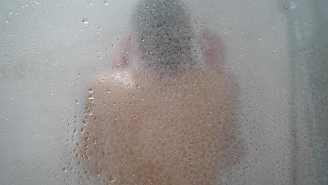A young woman is showering in a bathroom with a glass enclosure, the steamy water creates a misty and obscured view.
