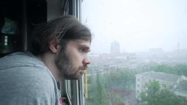 Depressed, sad man staying alone near window with pensive face. Downpour, heavy rain outside the window, bad weather. Sad, melancholy mood. Mental health concept. Sad man beard looking outside window