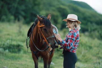 Obraz premium Happy blonde with horse in forest. Woman and a horse walking through the field during the day. Dressed in a plaid shirt and black leggings.