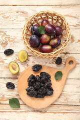Board with tasty dried plums and wicker bowl of fresh fruits on white wooden background