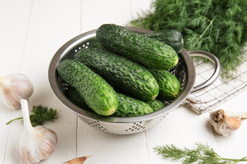 Colander with fresh cucumbers, garlic and dill on white wooden background
