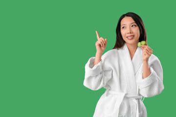 Young Asian woman with cucumber slices pointing at something on green background