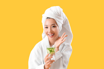 Young Asian woman with glass of cucumber water on yellow background