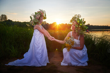 Mother and daughter in white sundresses, wreaths of flowers and bouquet on nature in evening at...