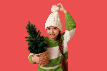 Cute little girl wearing warm hat with Christmas tree in pot on red background