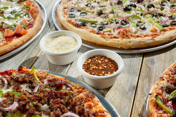 A view of condiment cups of parmesan cheese and red pepper flakes, surrounded by pizzas.