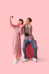 Couple of young tourists with suitcase taking selfie on pink background