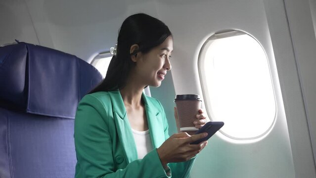 A young Asian businesswoman, dressed in a formal suit, sits at her airplane seat with a laptop and briefcase, utilizing in-flight Wi-Fi for a conference call.