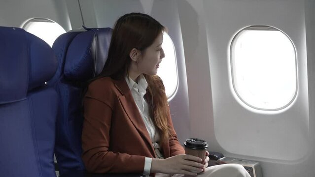 A young Asian businesswoman, dressed in a formal suit, sits at her airplane seat with a laptop and briefcase, utilizing in-flight Wi-Fi for a conference call.