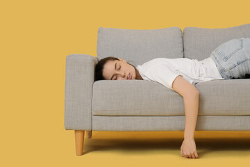 Tired young woman lying on grey sofa against yellow background