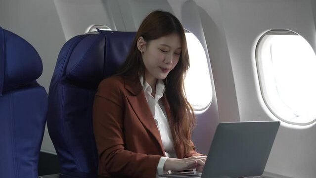 A young Asian businesswoman, dressed in a formal suit, sits at her airplane seat with a laptop and briefcase, utilizing in-flight Wi-Fi for a conference call.