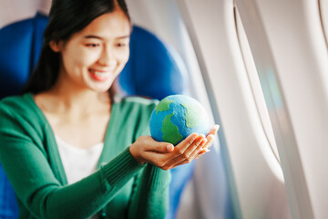 A young Asian woman, an airplane passenger, sits at her seat holding a world globe. Her travel essentials include a boarding pass, Earth day, Sustainable Fuel and Green energy, Save the world.